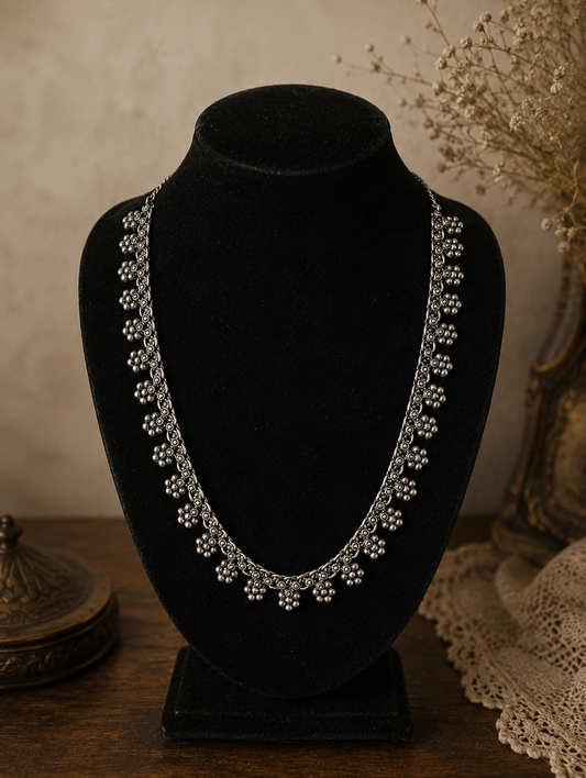 An antique-style oxidized silver necklace featuring intricate, repeating floral cluster charms, showcased on a black velvet bust display against a rustic backdrop.