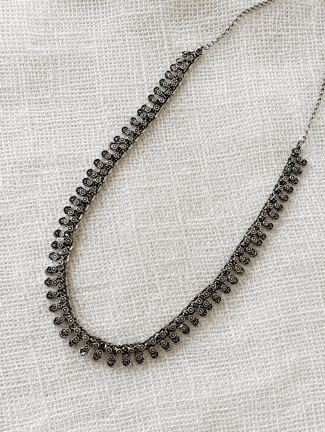 A delicate, antique-finish oxidized silver necklace featuring a continuous row of small, intricately detailed floral drop motifs, laid flat on a white textured fabric background.
