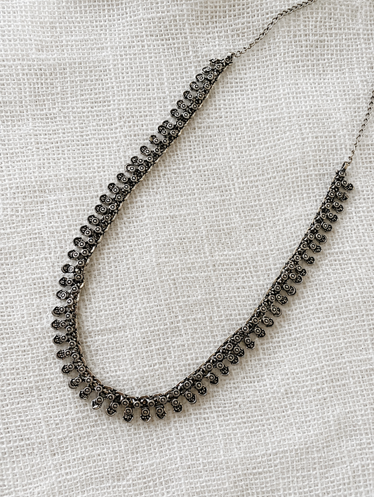 A delicate, antique-finish oxidized silver necklace featuring a continuous row of small, intricately detailed floral drop motifs, laid flat on a white textured fabric background.