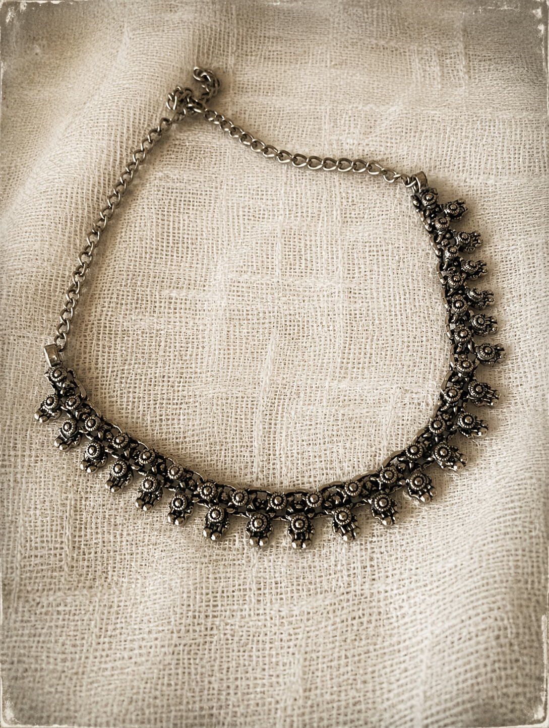 An antique-finish oxidized silver choker necklace featuring small, detailed geometric and floral fringe charms, arranged on a textured cream-colored fabric surface.