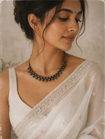 Woman wearing a white saree with intricate detailing and a black necklace against a neutral background