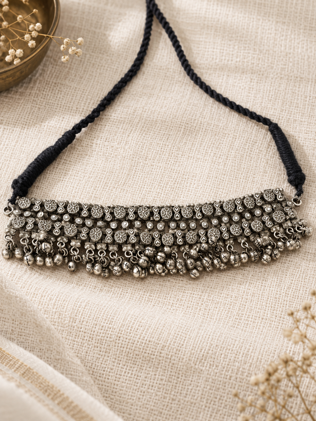 A bohemian-style silver bell necklace with a black braided cord, displayed on a textured beige linen fabric background with dried floral accents.