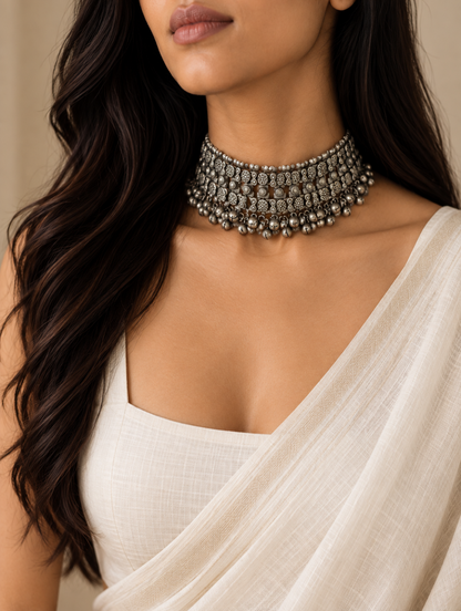 A close-up of a woman wearing a wide, ornate silver-toned beaded choker necklace with intricate circular patterns and dangling bead details, paired with a draped off-white blouse.