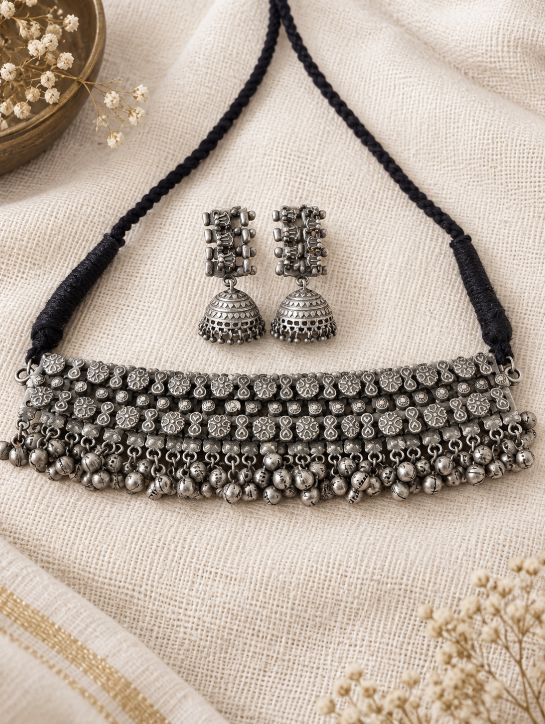 An antique silver-toned jewelry set featuring a multi-row necklace with a black braided cord and matching jhumka earrings, displayed on a textured beige linen fabric.