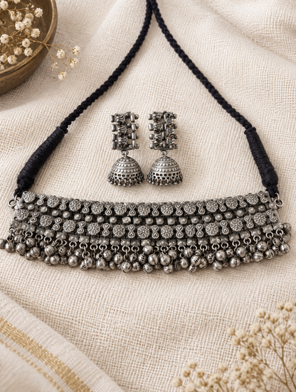 An antique silver-toned jewelry set featuring a multi-row necklace with a black braided cord and matching jhumka earrings, displayed on a textured beige linen fabric.
