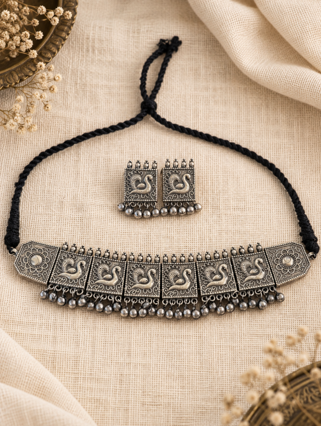 An antique silver-toned necklace with a matching pair of earrings and a black braided cord, displayed on a beige linen fabric background with dried floral accents.