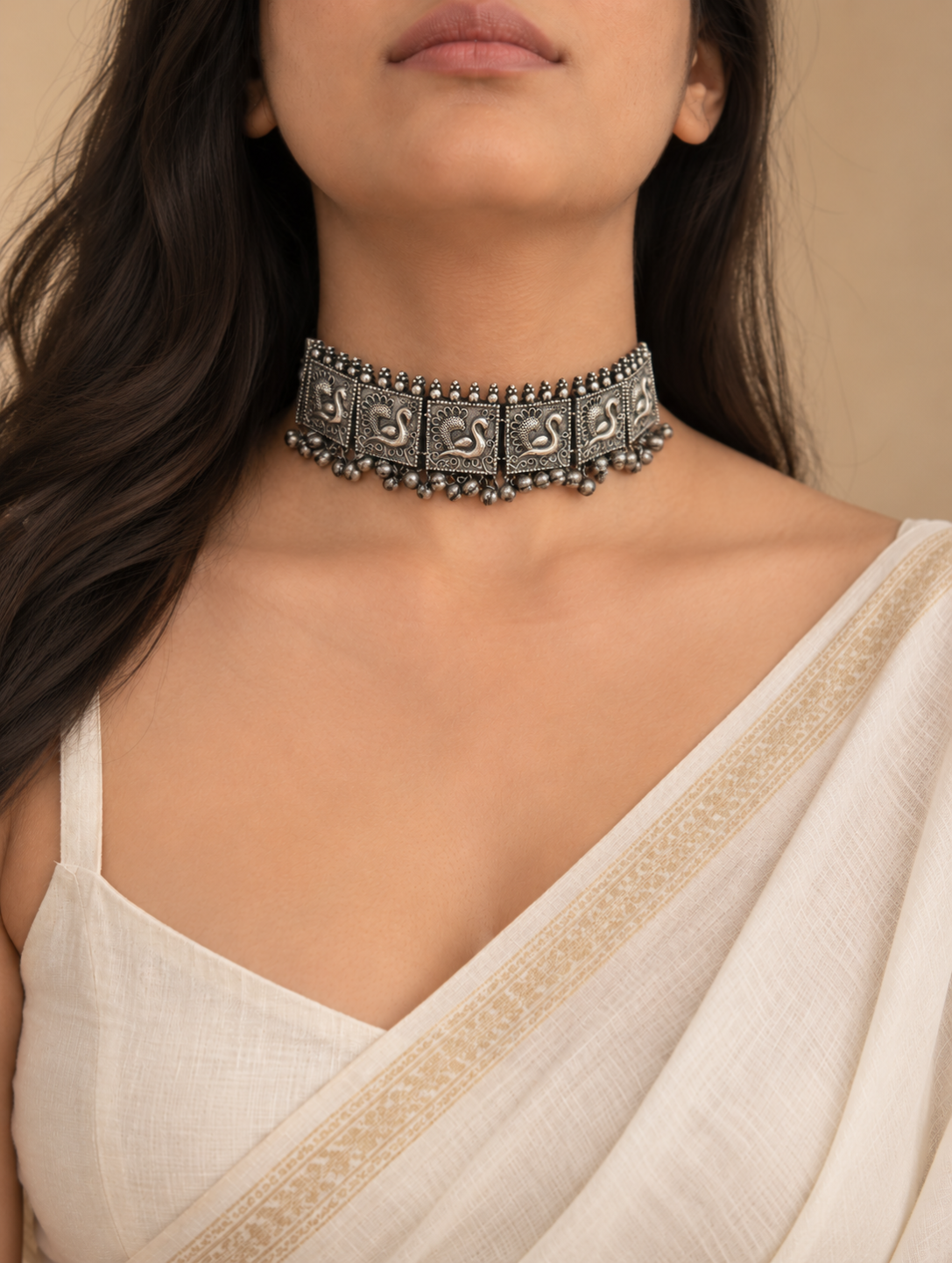 A close-up of a model wearing a wide, ornate silver-toned choker necklace with intricate engraved patterns and dangling bead details, paired with a cream-colored draped blouse.

