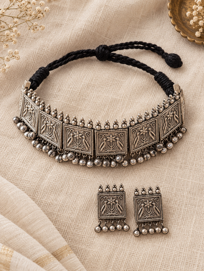An antique silver-toned choker necklace with matching rectangular earrings, displayed on a beige textured fabric background. The necklace features detailed embossed motifs on its silver panels, dangling silver beads, and a black adjustable braided cord.