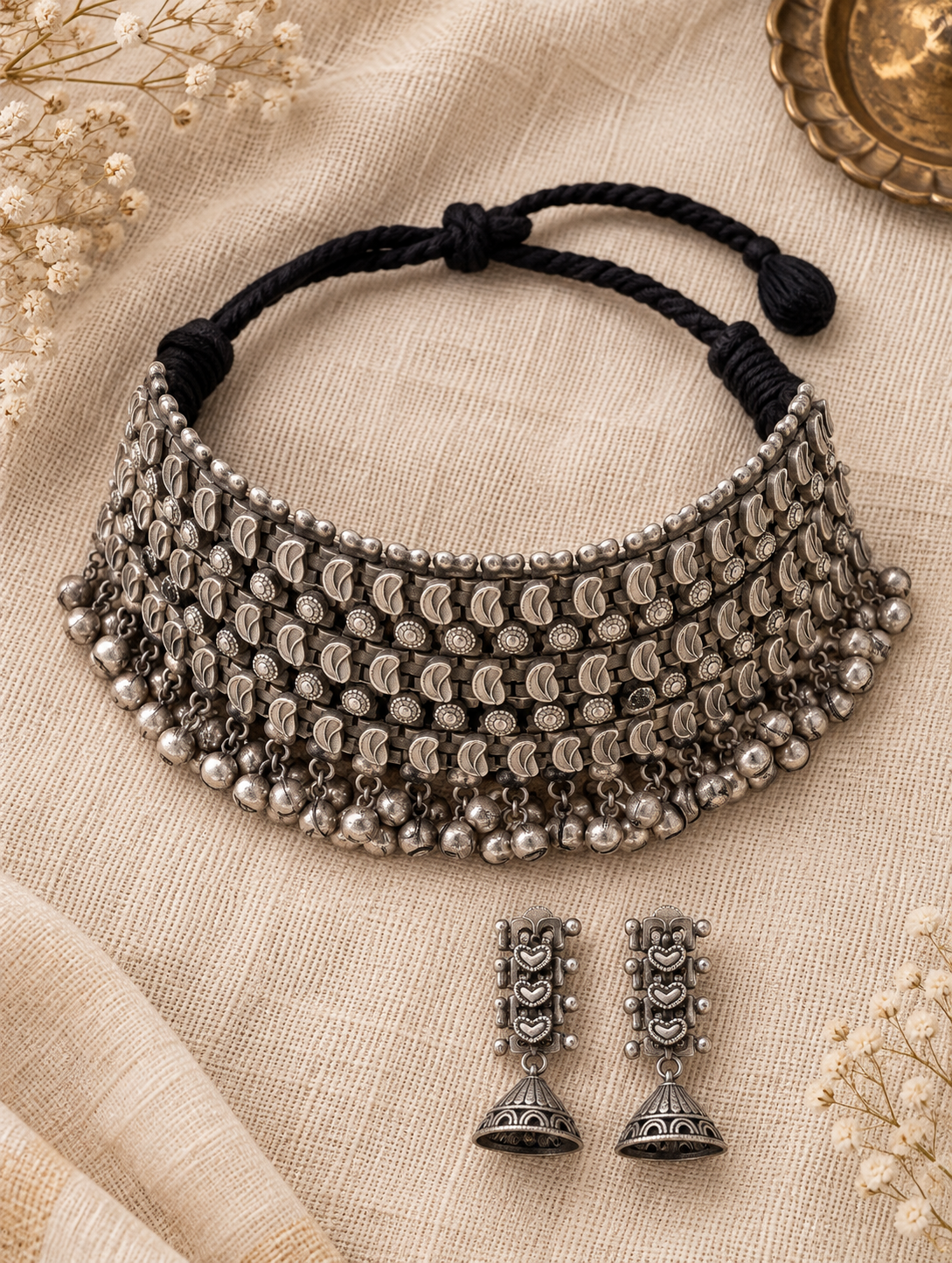 A silver-toned traditional Indian choker necklace with dangling jhumka-style bells and a matching pair of drop earrings, arranged on a beige fabric background with dried flowers and a brass plate.