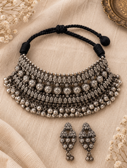 An ornate silver-toned traditional necklace with matching drop earrings, displayed on beige textured fabric with dried floral accents.