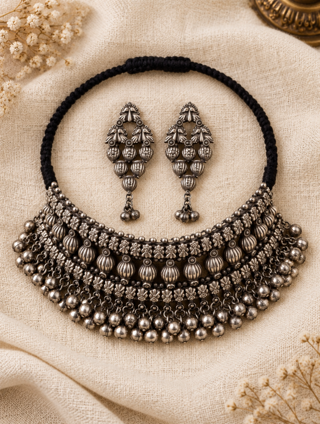 An ornate silver-toned statement necklace with matching drop earrings, displayed on a beige textured fabric background with dried floral accents.