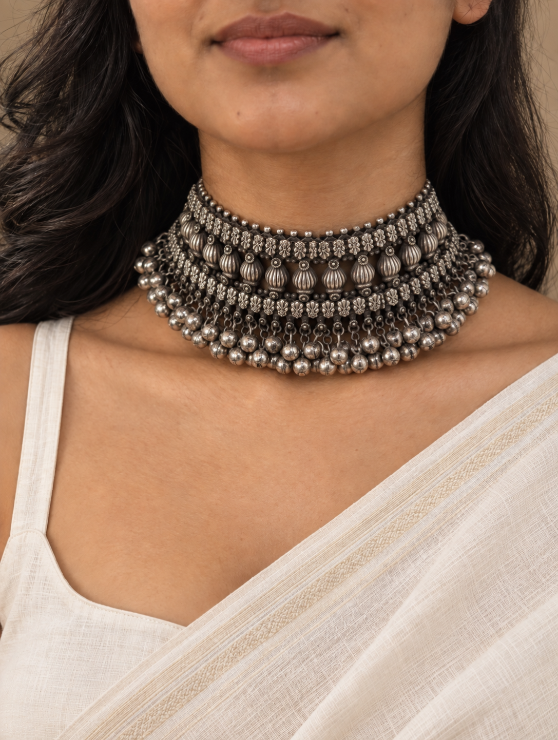 A close-up of a woman wearing a multi-strand silver-toned traditional choker necklace with intricate beadwork and dangling elements, paired with a white draped garment.