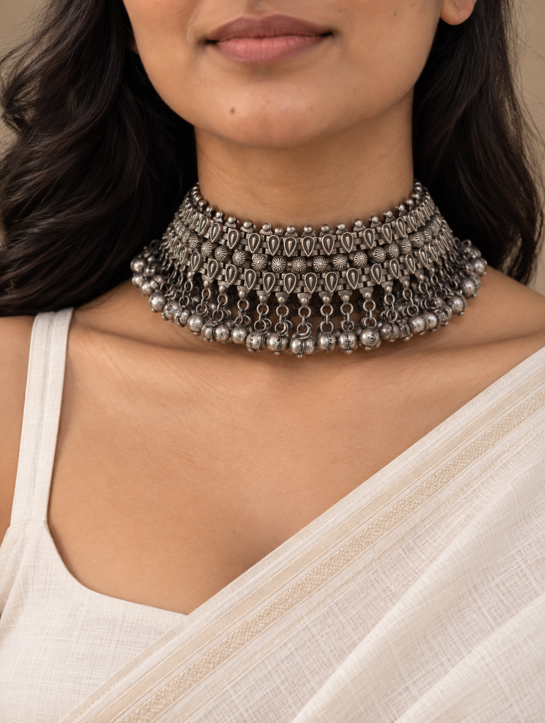 A close-up of a woman wearing a wide, ornate silver-toned choker necklace with intricate dangling links and bead details, paired with a light cream-colored top.