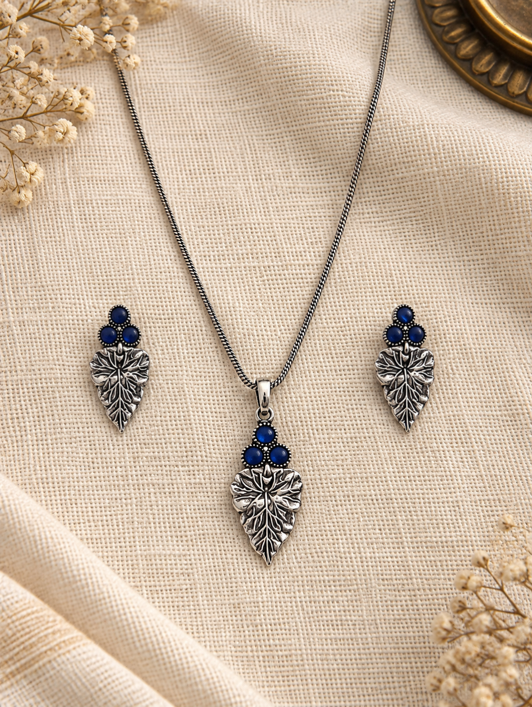 Silver-toned jewelry set featuring a necklace and matching earrings with leaf-shaped pendants and dark blue gemstone accents, arranged on a beige textured fabric background with dried floral decor.