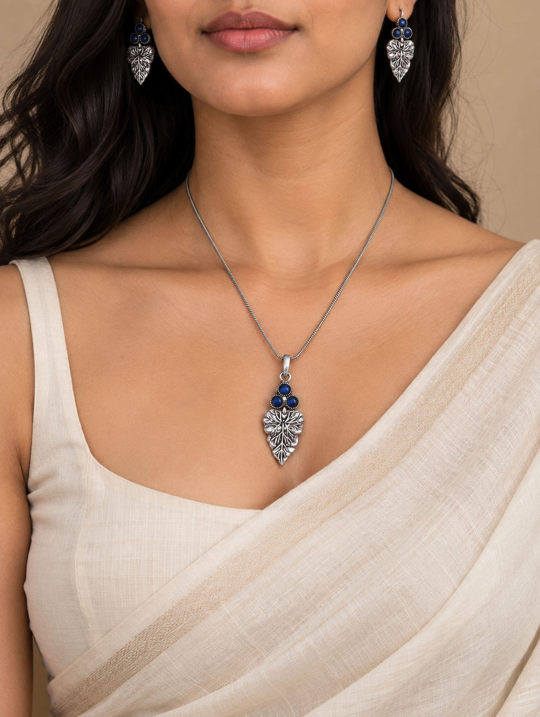 A close-up of a model wearing a silver-toned leaf-shaped pendant necklace with dark blue gemstone accents, matching drop earrings, and a cream-colored draped blouse against a warm beige background.