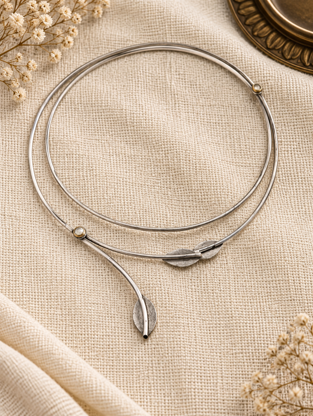A silver-toned bangle bracelet with a leaf-shaped charm and small bead details, displayed on a textured beige fabric with dried flowers.