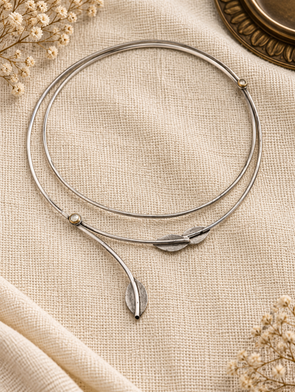 A silver-toned bangle bracelet with a leaf-shaped charm and small bead details, displayed on a textured beige fabric with dried flowers.