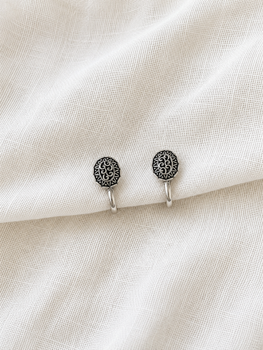A close-up product shot of two small, oval-shaped oxidized silver clip-on nose pins. Each nose pin is engraved with delicate filigree scroll patterns in the center, framed by a finely detailed tribal-style border, displayed resting against a soft, textured white fabric backdrop.