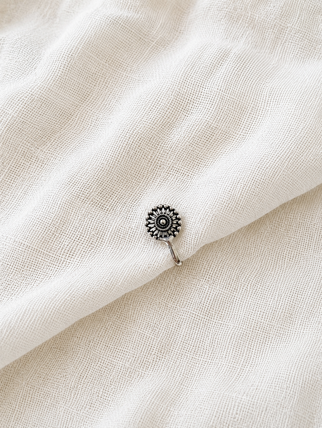 A high-angle product photograph showcasing a single, small oxidized silver clip-on nose pin. The nose pin is intricately designed in the shape of a miniature sunflower, featuring textured radiating petals around a raised center bead, resting elegantly on a soft, textured off-white fabric backdrop.