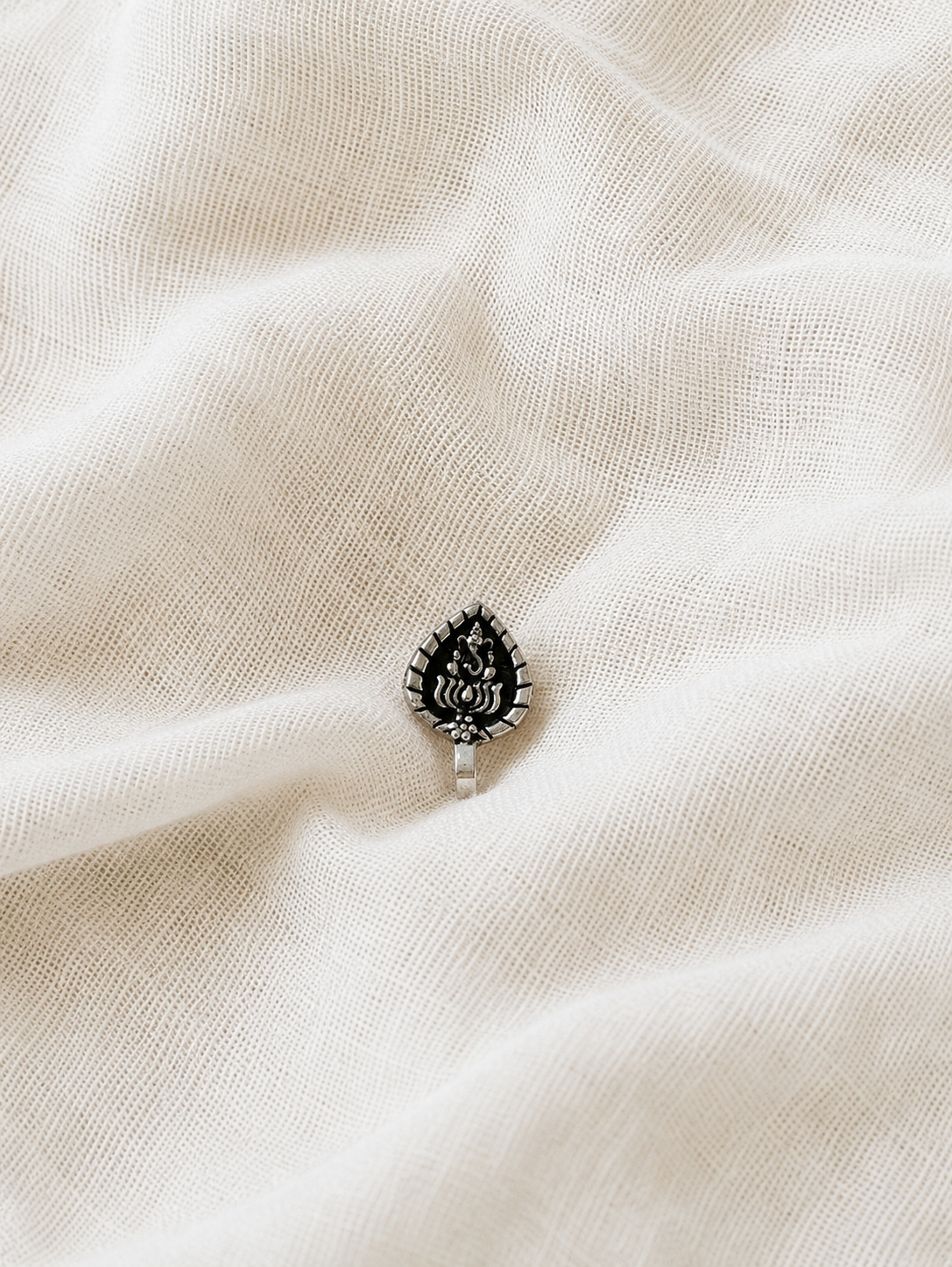 A high-angle product photograph showcasing a single, small clip-on nose pin made of oxidized silver. The nose pin is shaped like a pointed leaf or a betel leaf, featuring an intricately carved central motif of the Hindu deity Lord Ganesha seated on a lotus, contrasting beautifully against a darkened background and a textured white fabric backdrop.