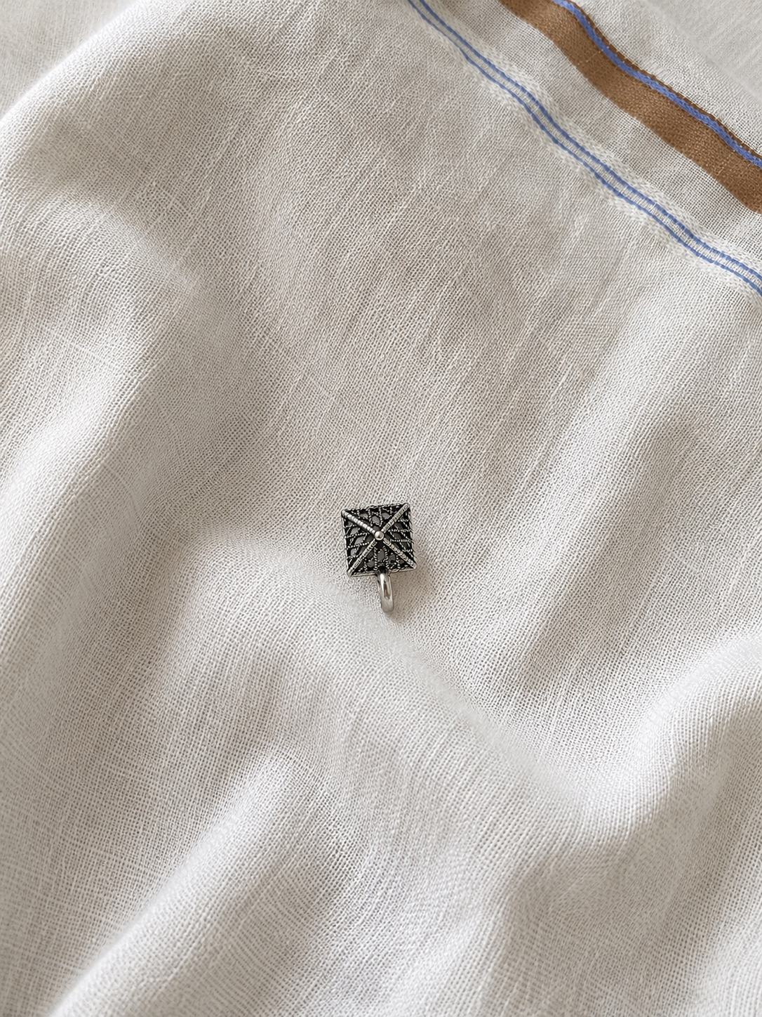 A high-angle product photograph showcasing a single, small clip-on nose pin made of oxidized silver. The piece is shaped in a perfect square, featuring an intricate geometric criss-cross lattice and openwork filigree design with a tiny central bead, resting flat against a soft, wrinkled white fabric background with a brown and blue bordered edge visible at the top.