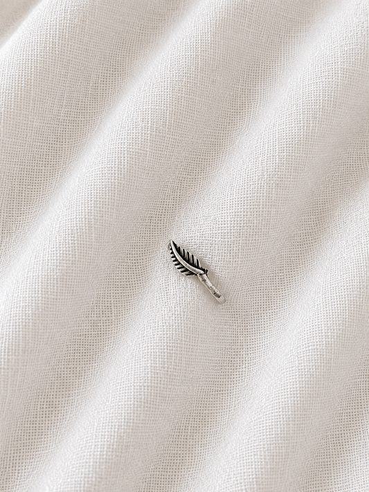 An image of a small, detailed silver nose pin shaped like a leaf resting on a textured, off-white fabric background, 