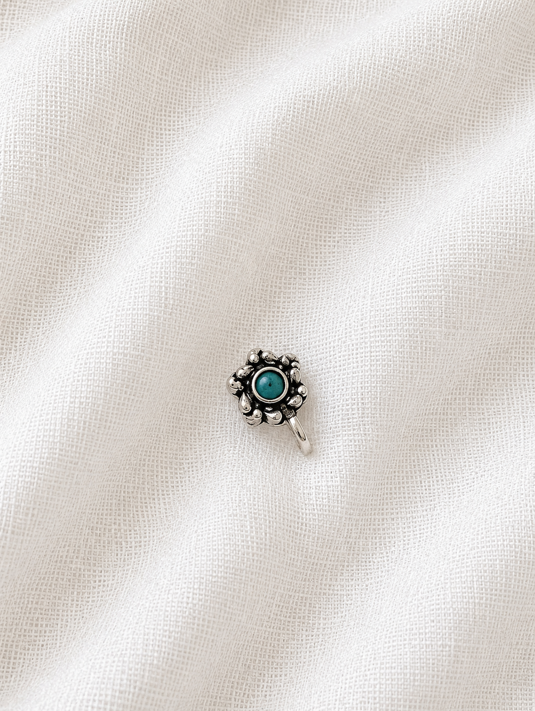 A small silver flower-shaped nose pin with a round turquoise gemstone center, showcased on a textured, off-white linen fabric background