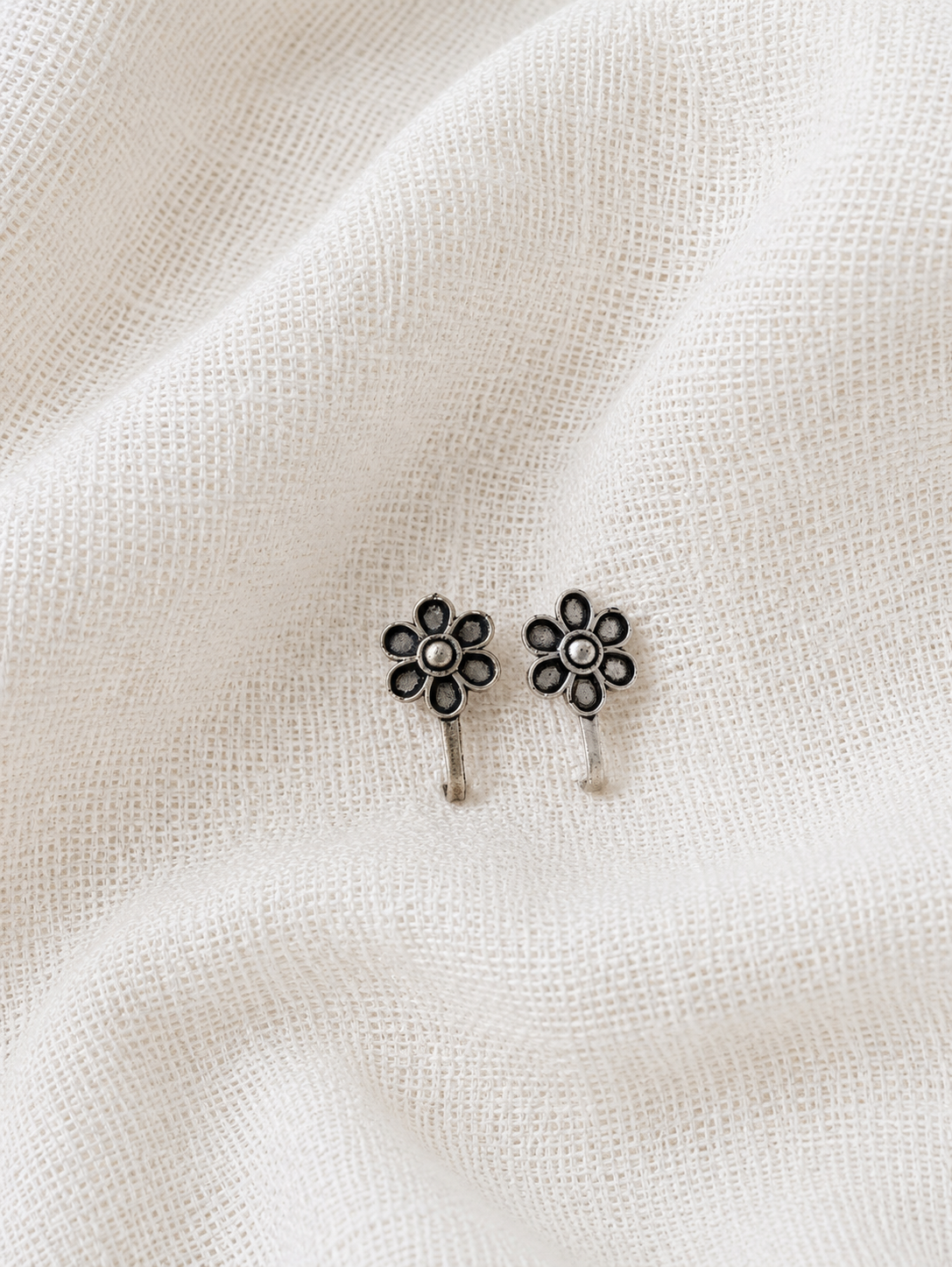 A pair of identical silver flower-shaped nose pins with dark oxidized detailing on the six petals, displayed side by side on a soft, wrinkled white linen fabric background