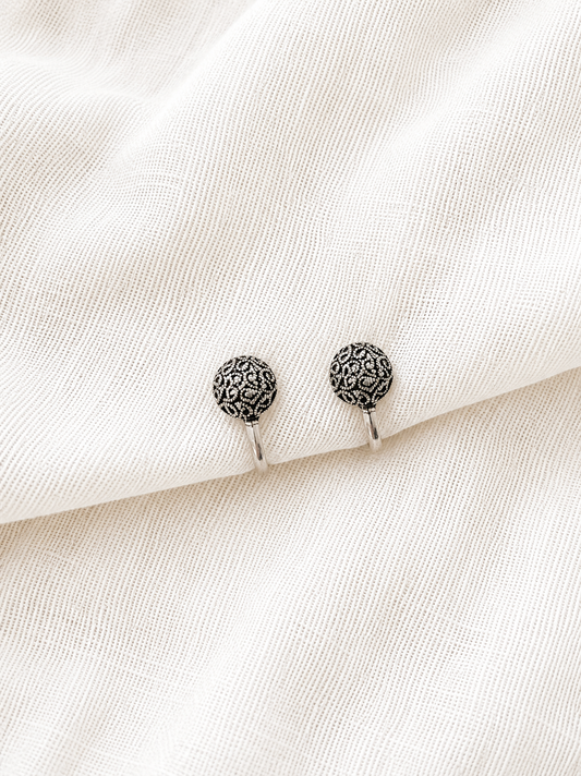 Two identical round, dome-shaped oxidized silver nose pins with intricate textured filigree patterns, resting side by side on a draped off-white woven fabric background 
