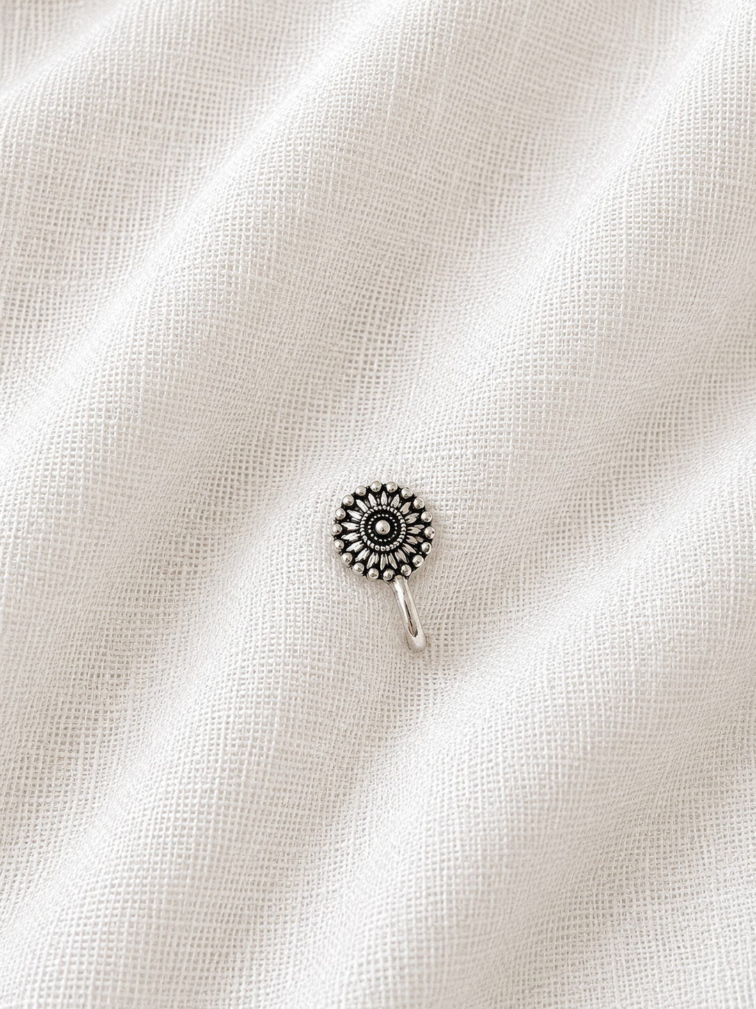 A single round, detailed silver nose pin shaped like a sunflower or mandala with intricate oxidized accents, resting on a textured, draped off-white linen fabric background 