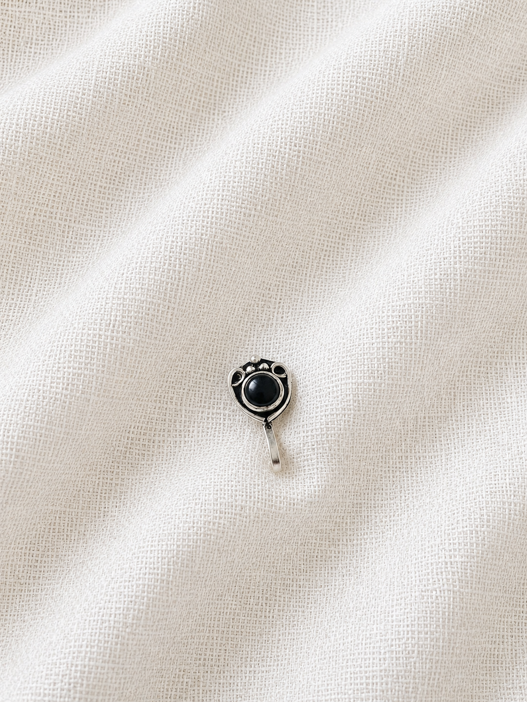 A small, round oxidized silver nose pin featuring a smooth black center stone with ornate silver framing and small bead details, displayed on a textured, off-white linen fabric background 