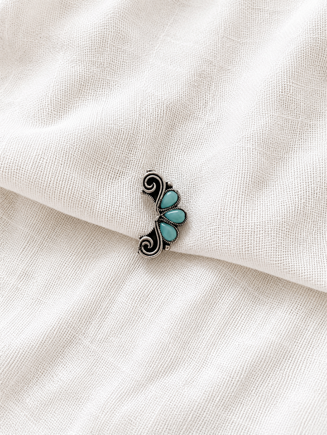 An oxidized silver half-moon crescent nath featuring three teardrop-shaped light turquoise stones and delicate filigree swirl borders, showcased on a folded off-white textured fabric background