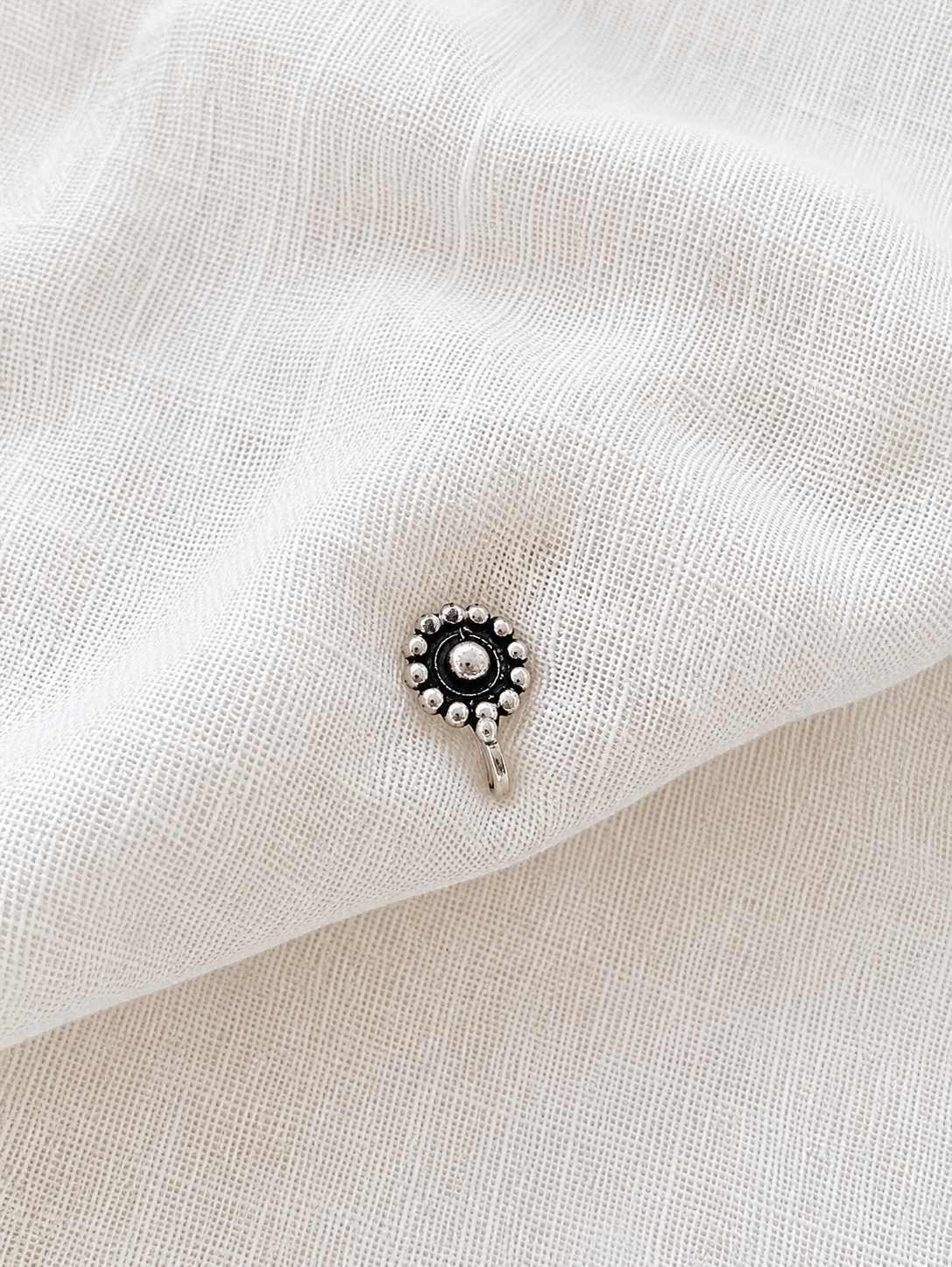 A small, round oxidized silver nose pin with a central metallic dome surrounded by a circular border of small silver beads, resting on a draped off-white textured fabric background 