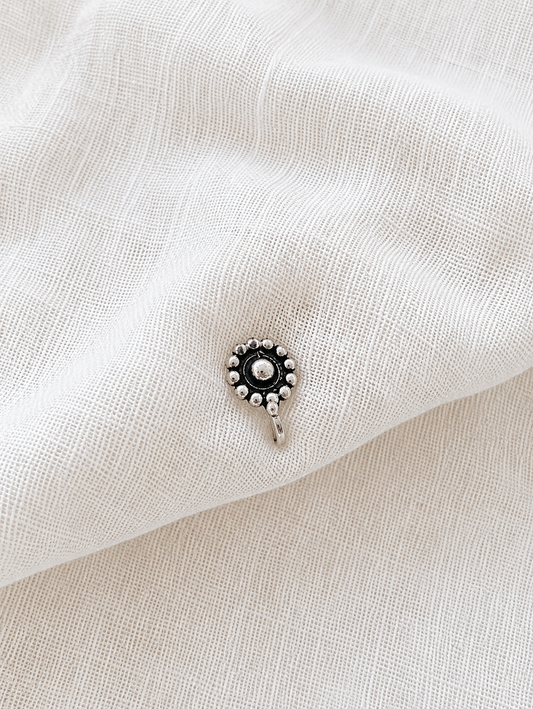 A small, round oxidized silver nose pin with a central metallic dome surrounded by a circular border of small silver beads, resting on a draped off-white textured fabric background 