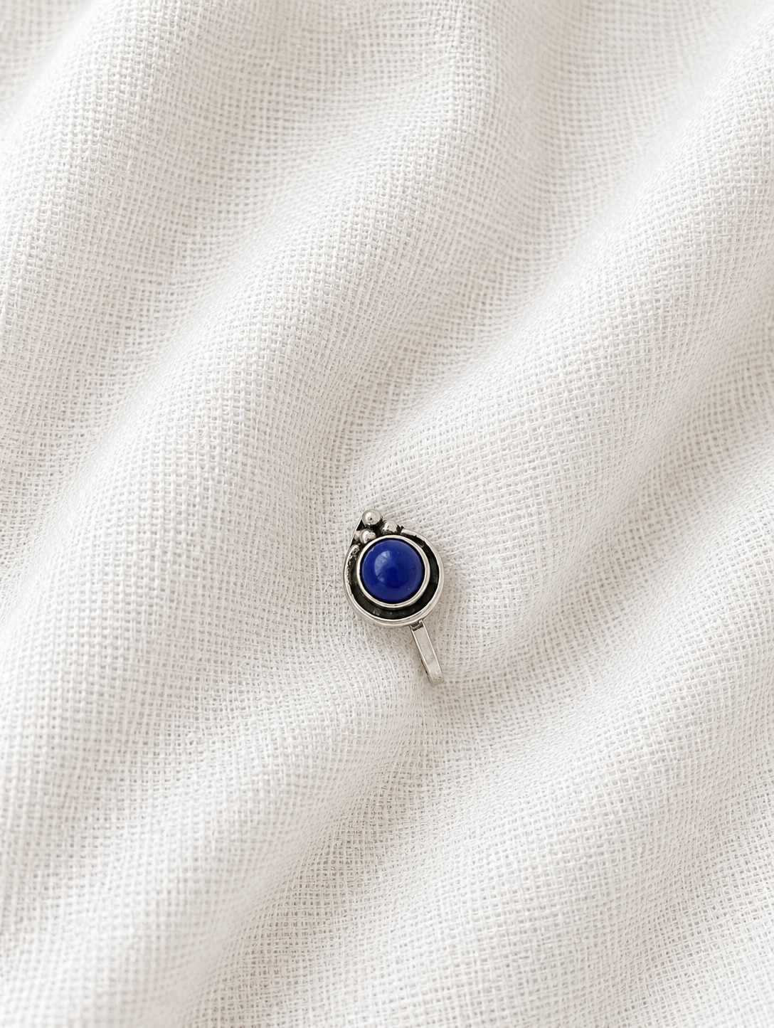 A small, circular oxidized silver nose pin featuring a smooth, vibrant blue round stone center with a tiny silver bead cluster at the top, resting on a draped off-white textured fabric background