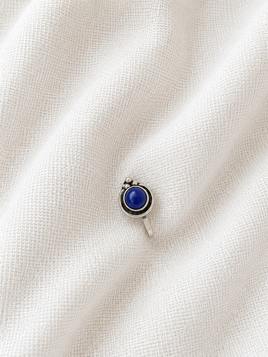 A small, circular oxidized silver nose pin featuring a smooth, vibrant blue round stone center with a tiny silver bead cluster at the top, resting on a draped off-white textured fabric background