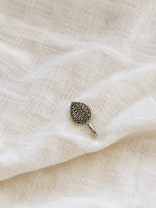 A small, leaf-shaped oxidized silver nose pin with a textured grid pattern of tiny silver lines and beads, resting on a draped, off-white textured fabric background