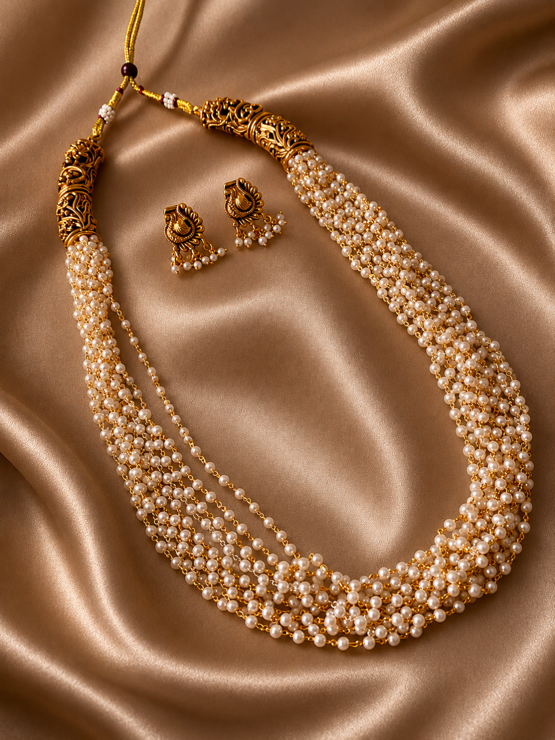 A traditional South Indian style multi-strand pearl long necklace and matching gold stud earrings with tiny hanging pearls, displayed on a black velvet bust against champagne silk.