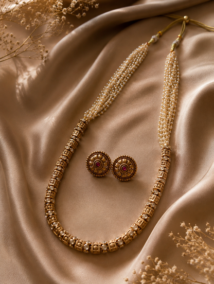 A classic South Indian long necklace featuring an antique gold block-patterned lower chain transitioning into multi-strand seed pearls, paired with matching round floral stud earrings on champagne silk fabric.