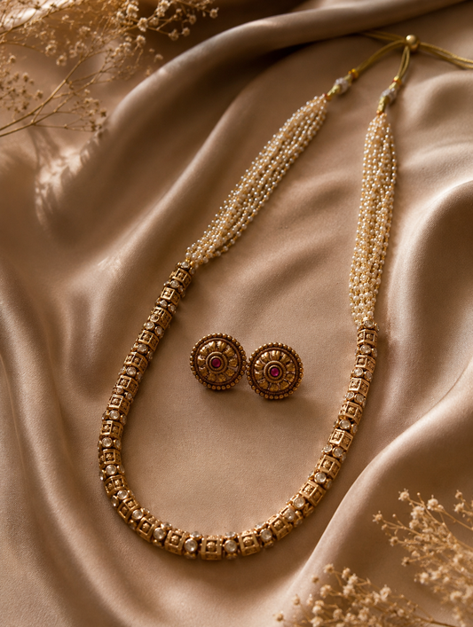 A classic South Indian long necklace featuring an antique gold block-patterned lower chain transitioning into multi-strand seed pearls, paired with matching round floral stud earrings on champagne silk fabric.
