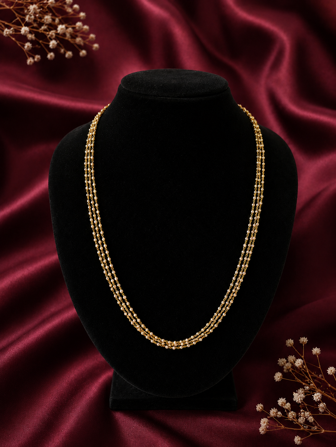 A dainty three-layered gold chain necklace featuring tiny woven seed pearls, displayed beautifully on a black velvet bust mannequin against a maroon satin background.