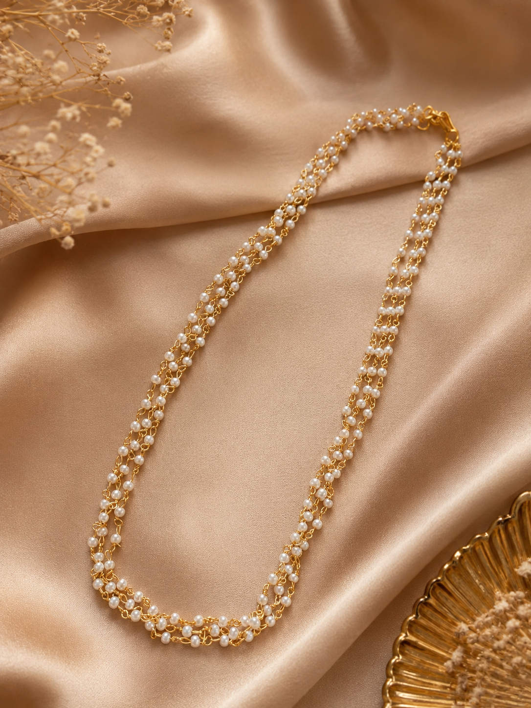 A delicate two-layered gold chain necklace interspersed with small, fine white pearls, arranged gracefully over a shimmering champagne-colored silk fabric backdrop.