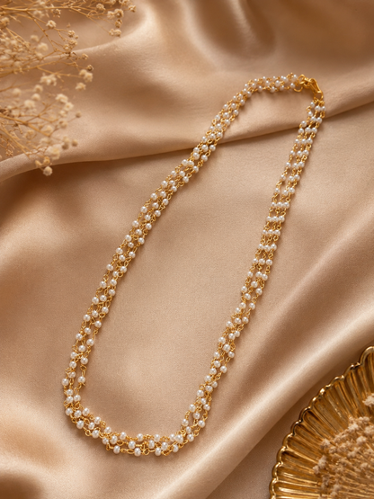 A delicate two-layered gold chain necklace interspersed with small, fine white pearls, arranged gracefully over a shimmering champagne-colored silk fabric backdrop.