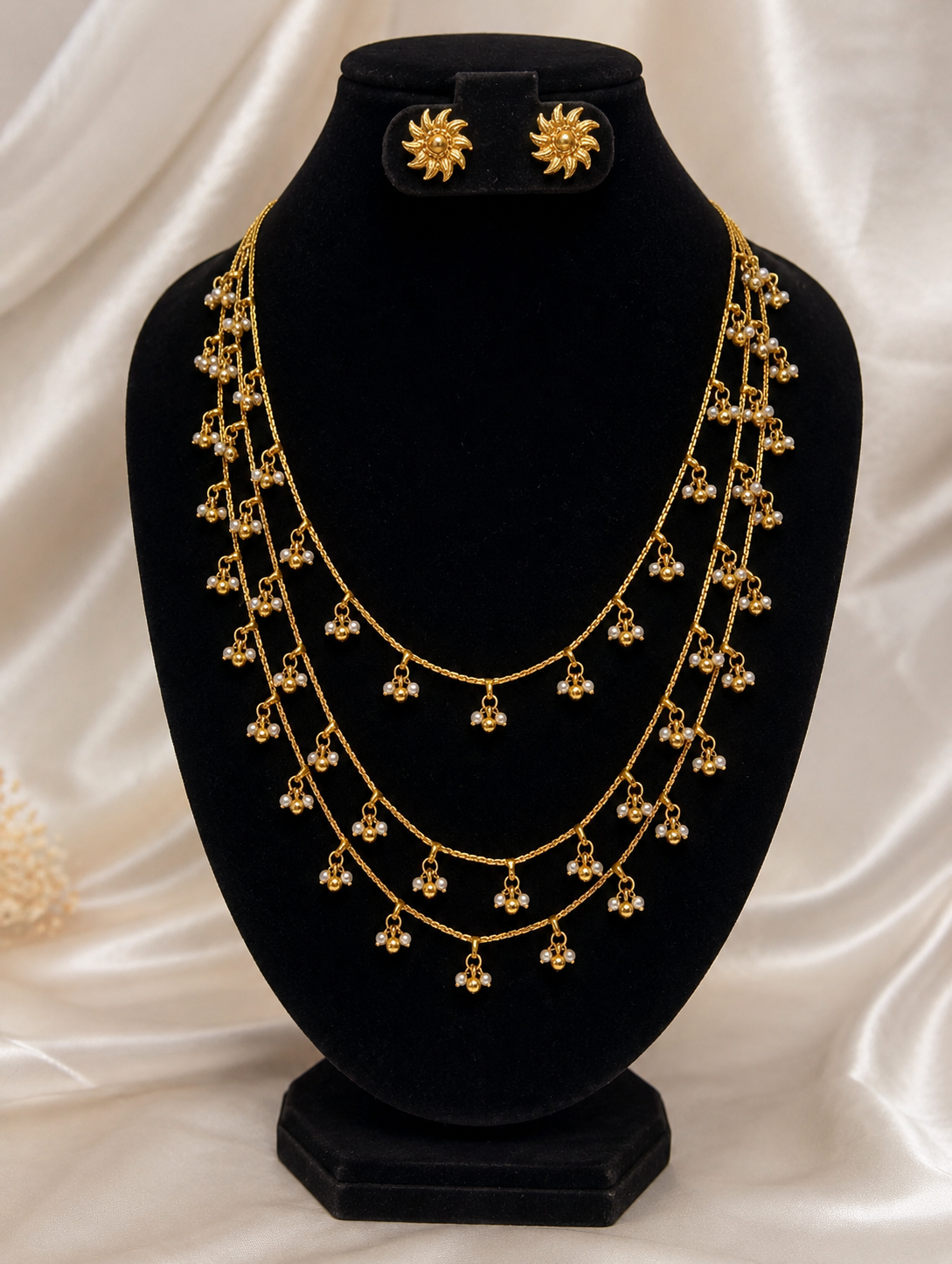 Traditional Shyamala jewelry set featuring a three-layered gold chain necklace with hanging tiny pearl droplets, paired with matching gold sunburst stud earrings, displayed on a black velvet bust.