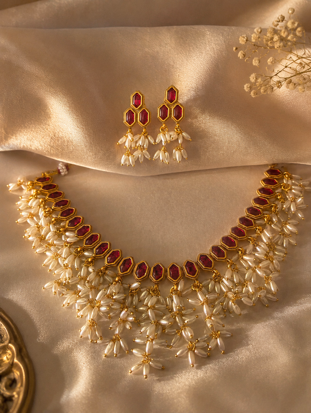 Traditional Rohini jewelry set featuring a gold necklace and matching drop earrings set with geometric red stones and dense bunches of white seed pearls, displayed on a shimmering champagne satin fabric.