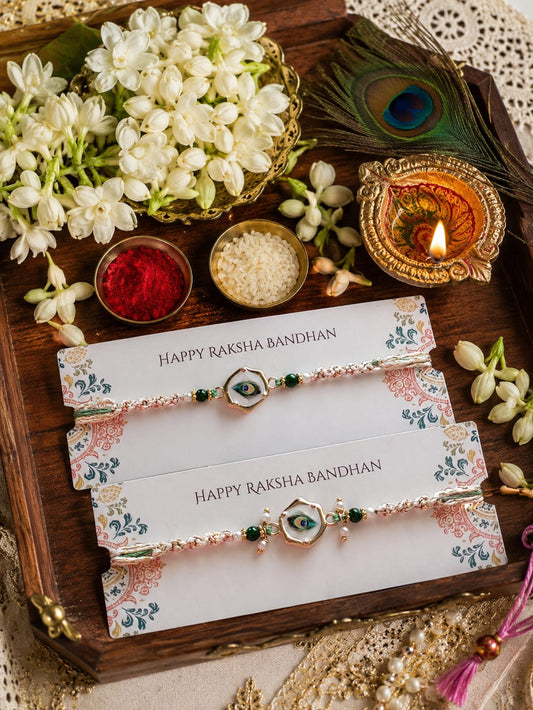 Three Rakhi bracelets wrapped in floral paper are arranged on a wooden tray with white flowers, a lit diya, peacock feathers, and small bowls of red and white powder. The paper labels read “HAPPY RAKSHA BANDHAN.