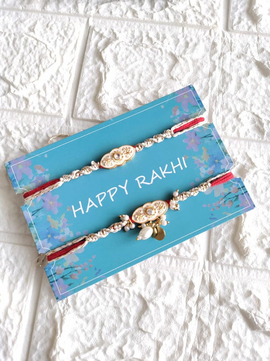 Two handcrafted designer Rakhis featuring oval ivory Meenakari charms with gold accents, dangling white pearls, and braided threads on a blue card.