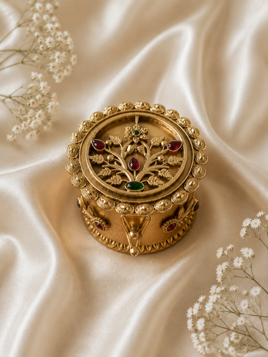 Ornate circular jewelry box made of antique gold-plated metal, featuring a domed lid with a sculpted tree of life motif adorned with delicate leaves and teardrop-shaped ruby and emerald stones. The box is decorated with textured bead trim and stands out beautifully against a soft, wrinkled cream satin backdrop.