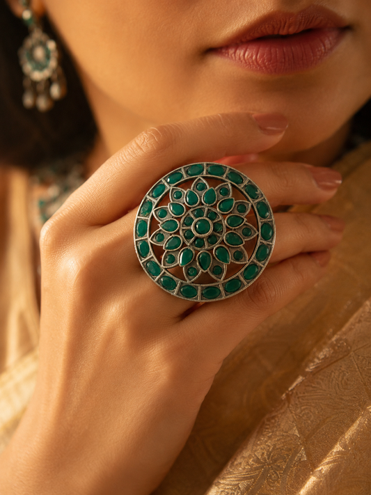 A close-up of a woman's hand wearing a large, circular oxidized silver statement ring with an intricate floral pattern inlaid with green stones.