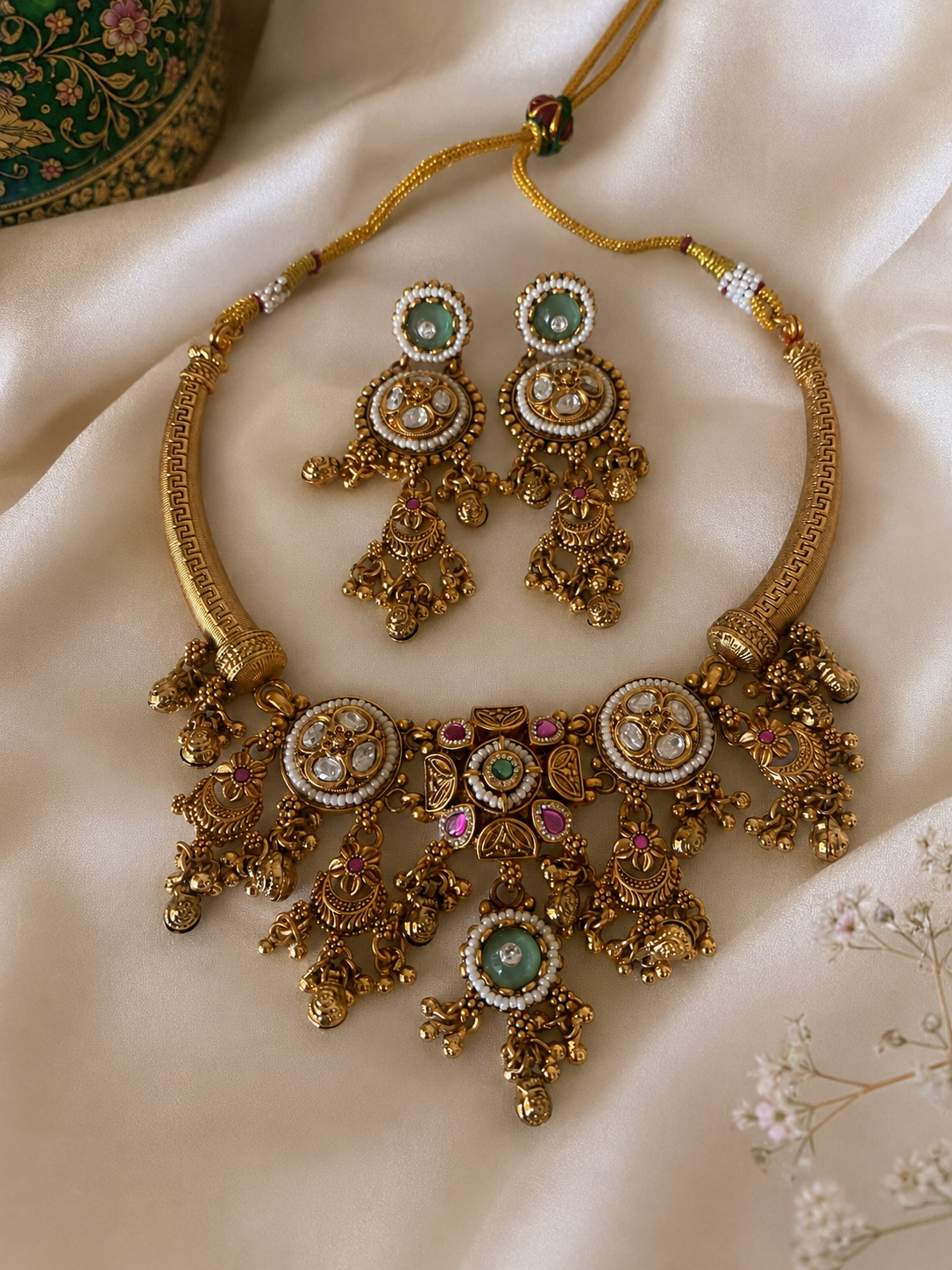 Rithwika traditional jewelry set displaying an antique gold-finished Hasli rigid collar necklace featuring round crystal polki medallions, a central cascading fringe with filigree gold drops, pink accent stones, and mint green emeralds, laid flat on soft cream satin fabric alongside matching long drop earrings.
